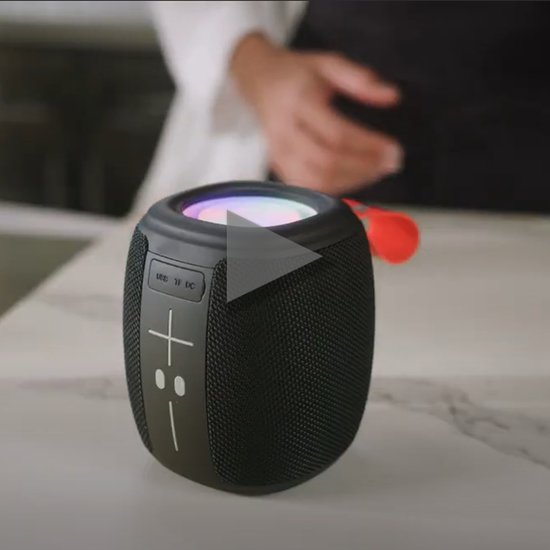 Video showing a woman in a kitchen - she places a small portable speaker on the counter, plays music and dances. Then cuts to another scene placing she speaking down by the strap